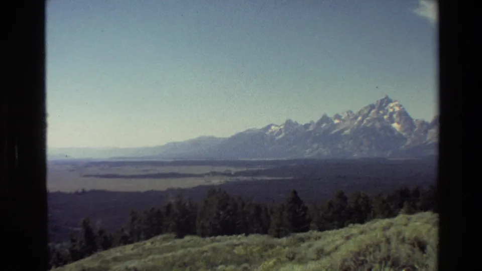 1980 8mm frame of the Grand Teton range with prominent snow-capped peaks, lake water, valley flats, and trees in the foreground.
