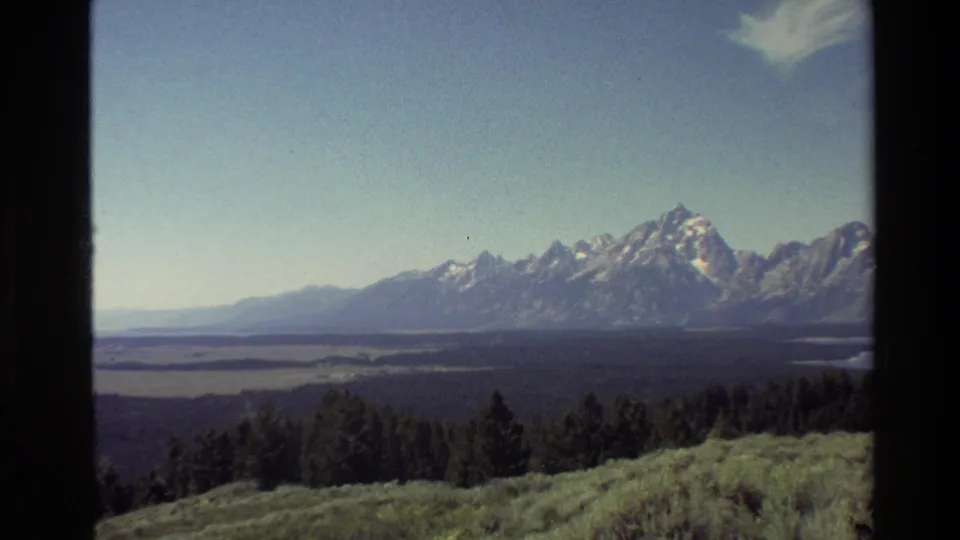 Grand Teton area overlook in 1980 8mm footage, showing a broad lake, wooded foreground, snow-capped range, and visible film-frame edges.