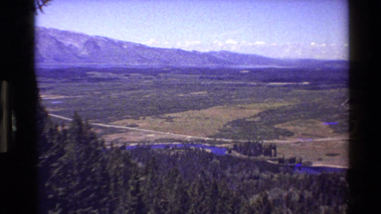 Wide mountain valley seen from above, with forested foreground, river, road, and distant peaks.