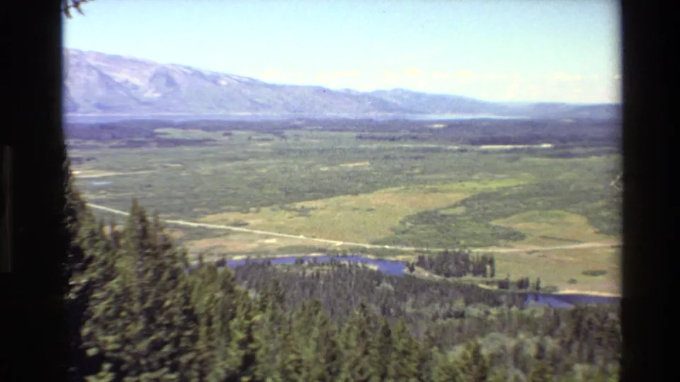 1980 home-movie view across Grand Teton valley terrain with evergreen foreground, open fields, river, road, and mountain ridge.