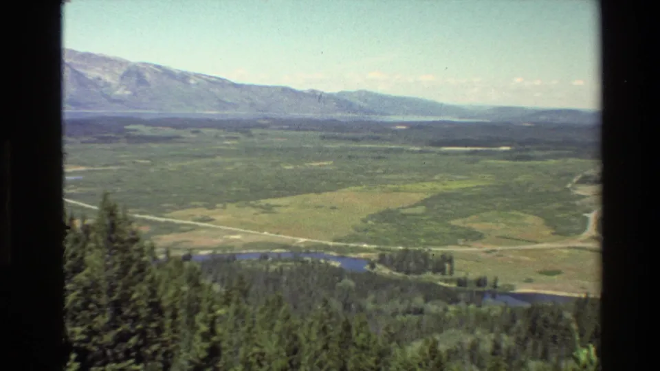 Grand Teton landscape view on 8mm film, with forest in the foreground and a wide valley, river, road, and mountains beyond.