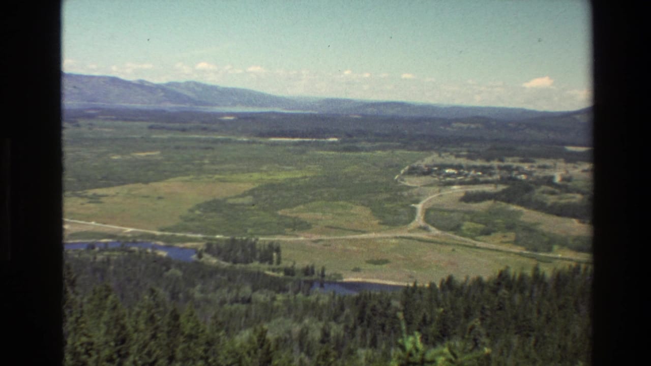 Wide valley landscape with mountains, forest bands, roads, water, and small buildings.