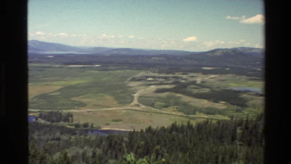 1980 Grand Teton home-movie view across open fields and forest toward distant hills, with a curving road and water visible below.