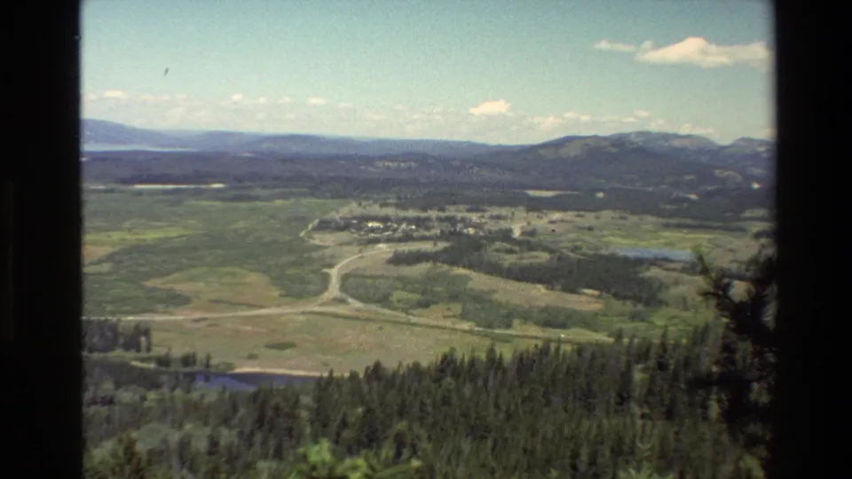 1980 Grand Teton frame with forested foreground, broad valley, roads, small settlement, water, and distant mountain ridges under blue sky.
