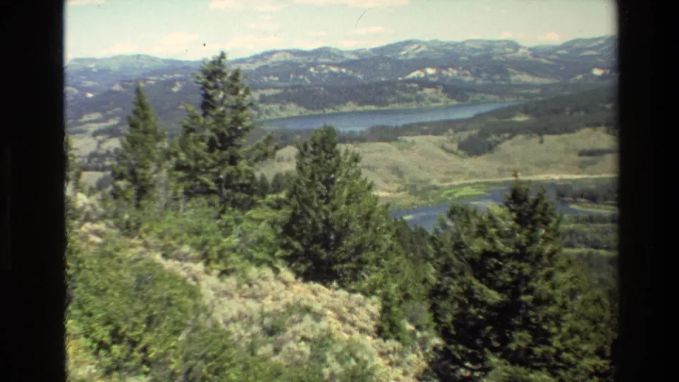 1980 8mm Grand Teton home-movie frame with evergreens in the foreground and a broad valley, water, and mountain ridges beyond.