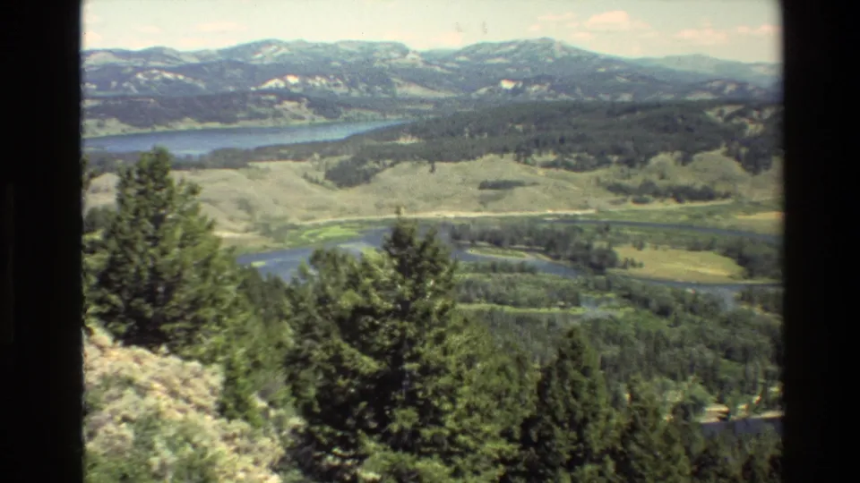 1980 Grand Teton, Wyoming frame with foreground conifers, open valley, blue lake water, and distant mountain ridges.