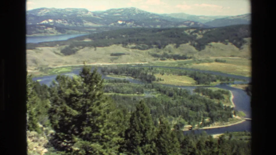 1980 Grand Teton, Wyoming frame showing a river bend below a forested overlook, with lake water and mountains in the distance.