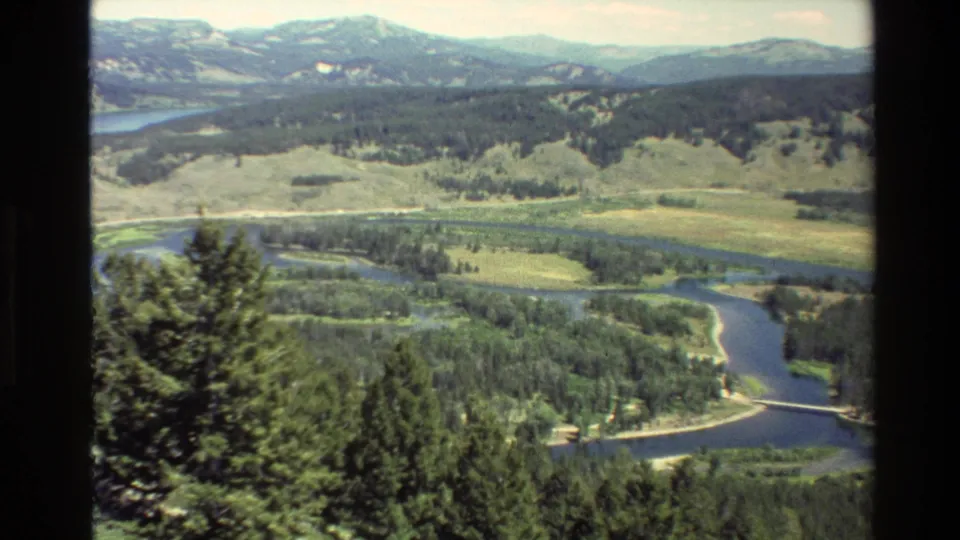 Grand Teton, Wyoming landscape with blue river channels, green forest, pale meadow, and distant ridges in a vintage 8mm frame.