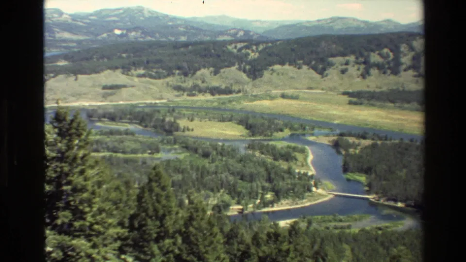 1980 8mm view over a Grand Teton river bend with dense trees, open meadow, low hills, and a small bridge or crossing near the water.