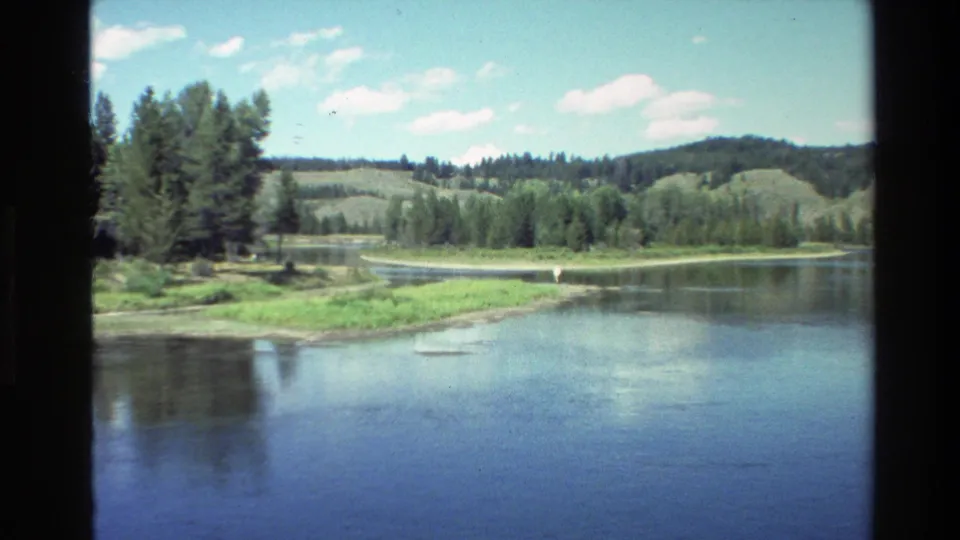 Wide Grand Teton landscape view shows still water, tree-lined banks, low hills, and scattered clouds in 1980 home-movie footage.