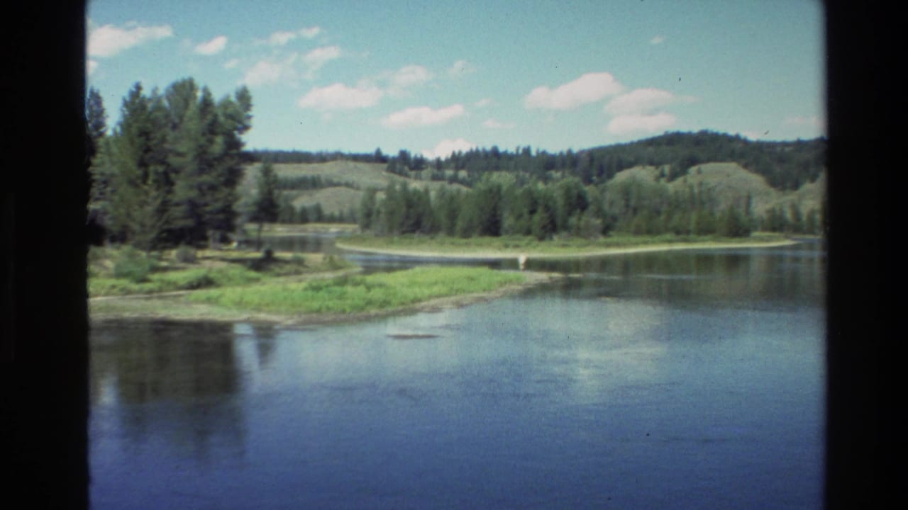 Calm lake or river bordered by trees and hills under a bright sky.
