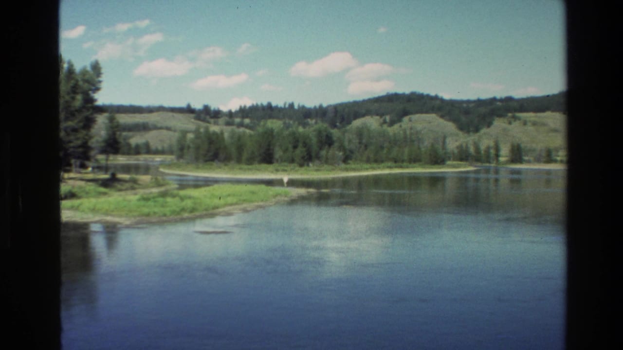 Wide water view with trees, grassy banks, hills, and clouds.