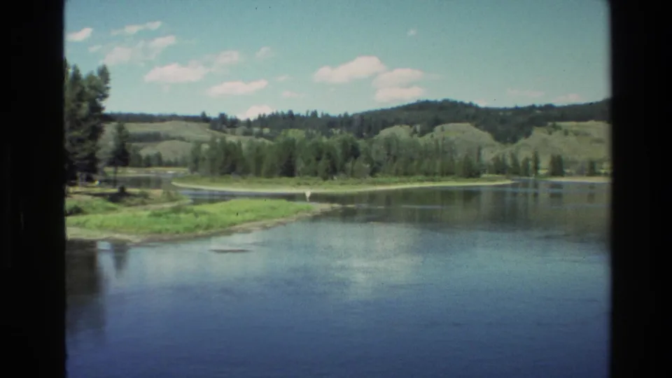 Grand Teton, Wyoming home-movie frame showing a quiet lake or river with wooded banks and low hills in sunny weather.