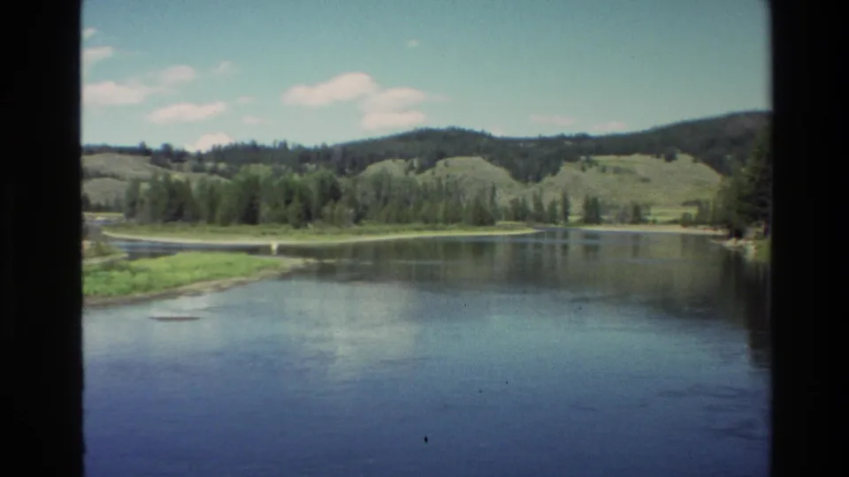 Vintage 8mm frame from the Grand Teton area showing reflective water, a low green bank, trees, and hills beyond.