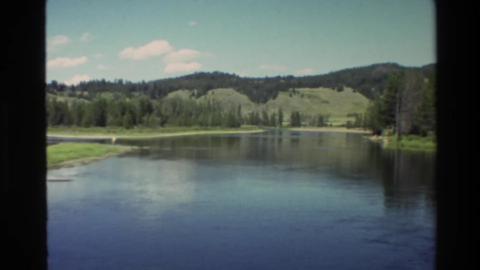 1980 Grand Teton area landscape frame with calm water, trees on the far bank, rolling hills, and a bright partly cloudy sky.