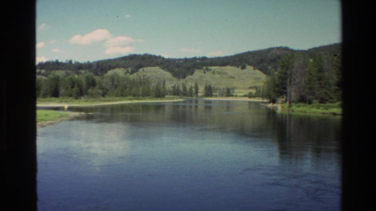 1980 Grand Teton, Wyoming home-movie view of calm water, pine-lined banks, and low green hills framed by dark film edges.