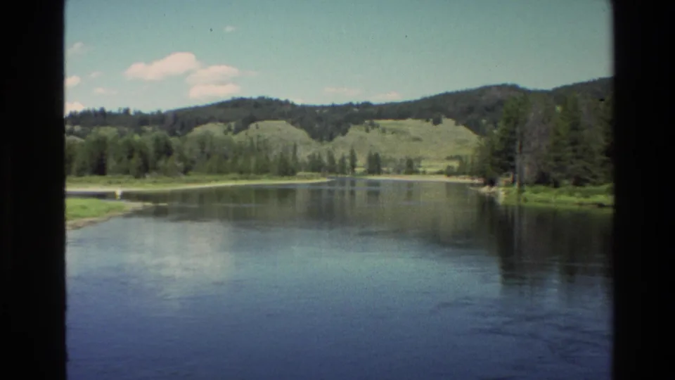 1980 Grand Teton, Wyoming home-movie view of calm water, pine-lined banks, and low green hills framed by dark film edges.