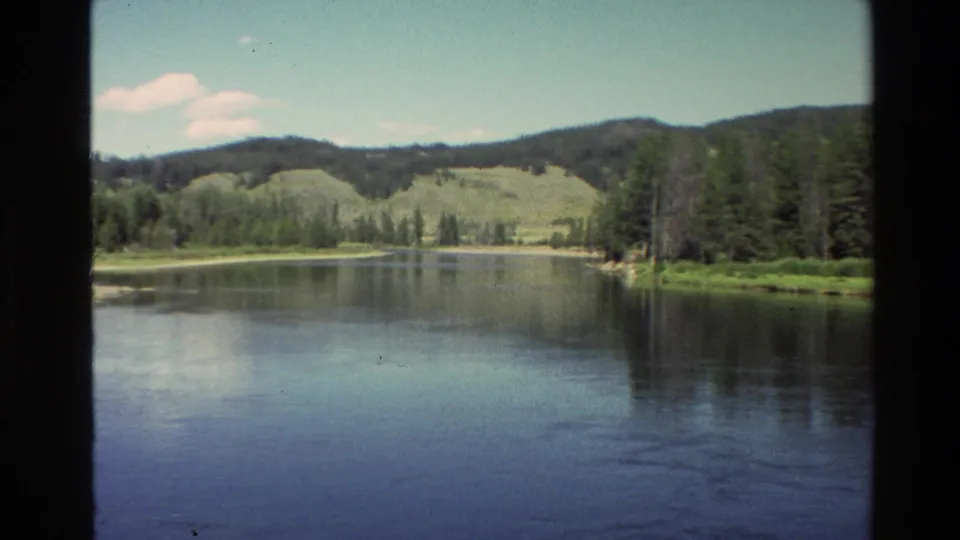 Grand Teton-area river landscape on 8mm film, showing still water, pine forest, grassy hills, and a shaded window-like border.