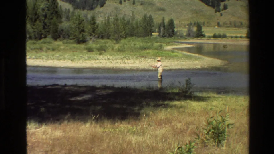 A lone fisherman in waders and a hat stands in the river near a grassy bank in 1980 Grand Teton, Wyoming home-movie footage.