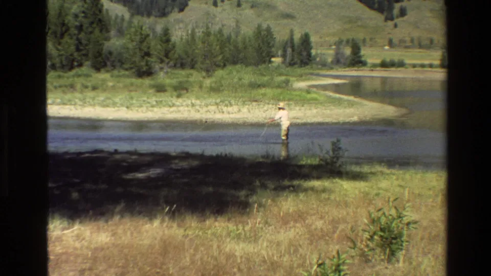 Distant fisherman in a bright shirt stands near the riverbank, with reeds, pine trees, and hills visible in the Grand Teton-area landscape.