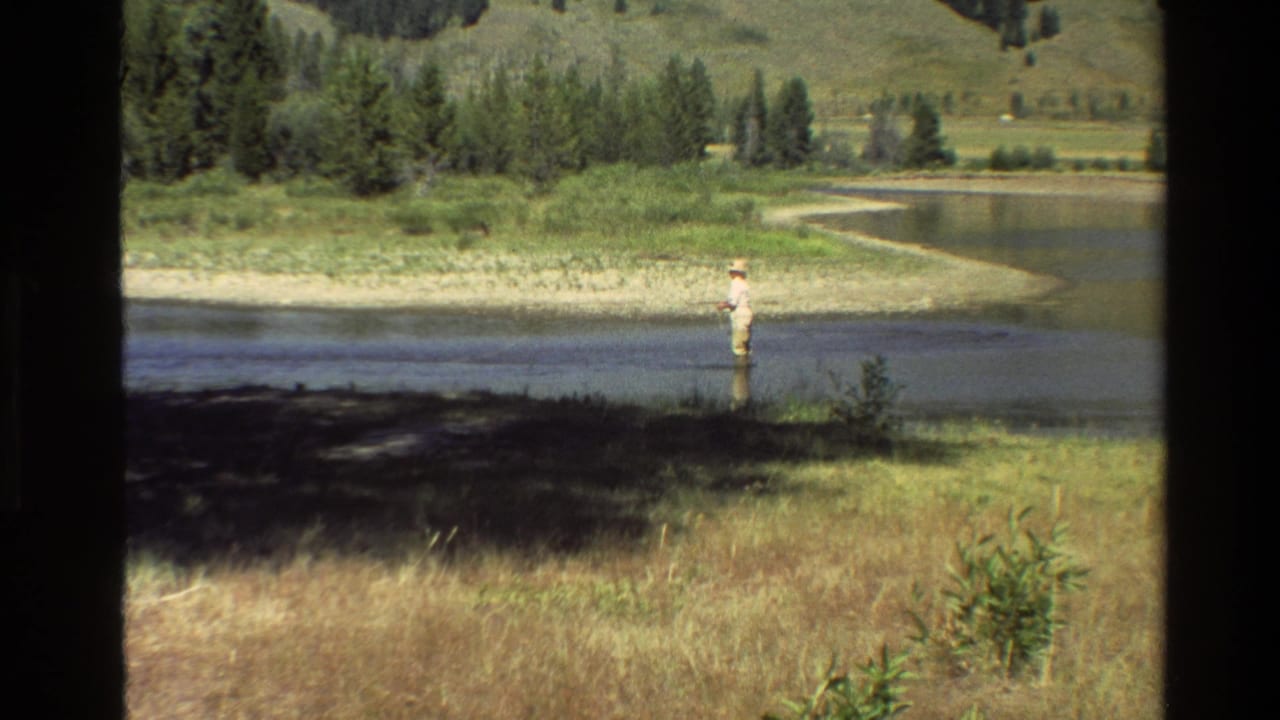 1980 Grand Teton home-movie frame showing a distant man fly fishing in a quiet stream bordered by grass, trees, and rolling hills.