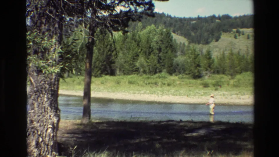 1980 Grand Teton home-movie still of a distant angler in a stream, framed by dark shade, tree trunks, grass, and forested hills.