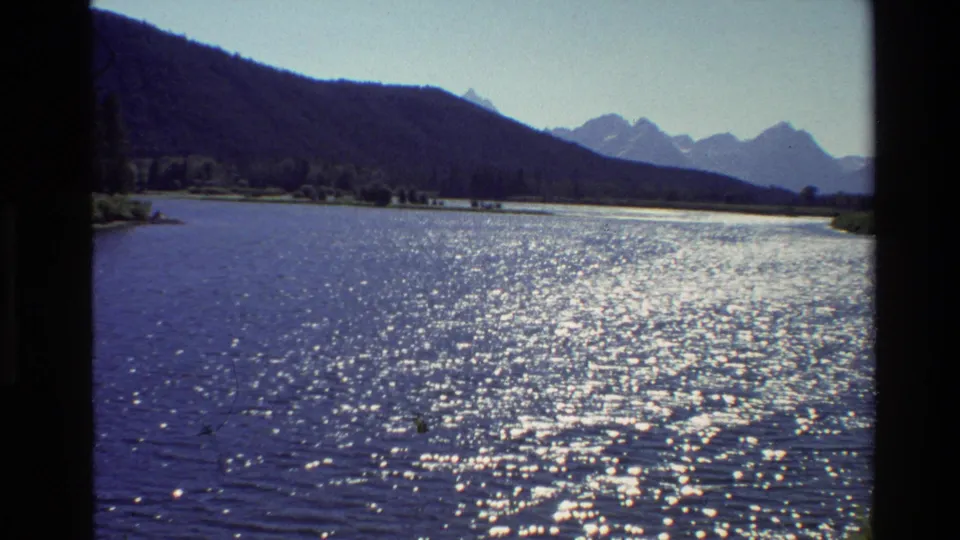 1980 Grand Teton home-movie frame showing a broad lake surface, bright reflected sunlight, dark tree line, and layered mountain ridges.