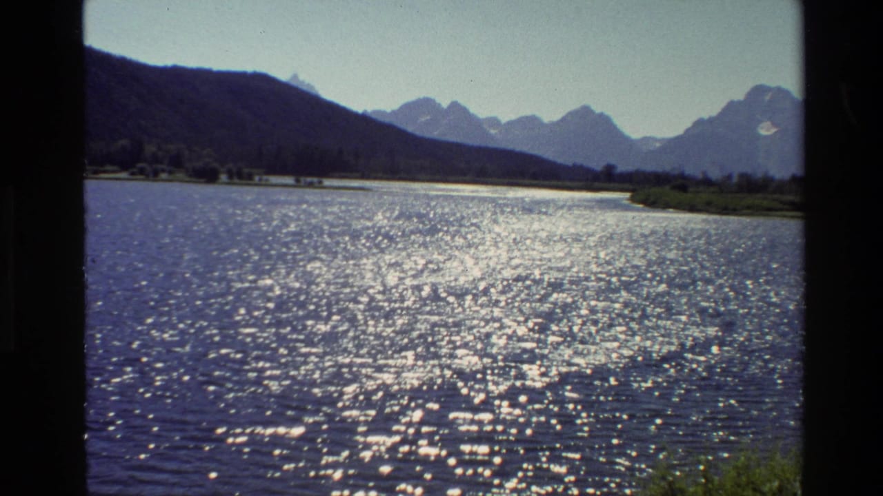 1980 Grand Teton, Wyoming 8mm frame showing sunlit lake water, distant mountains, green shore, and blue sky within dark film borders.