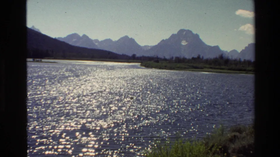 1980 8mm Grand Teton scenic frame with sparkling lake water, grassy foreground, distant mountains, and small clouds over a blue sky.