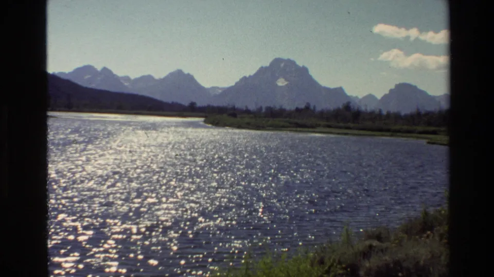 Grand Teton, Wyoming 1980 home-movie frame showing a wide sunlit lake, green banks, distant mountains, scattered clouds, and dark 8mm borders.