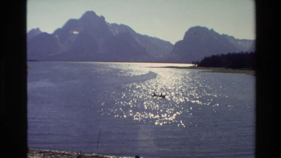 1980 Grand Teton footage with a small distant boat on reflective water and mountains in the background.