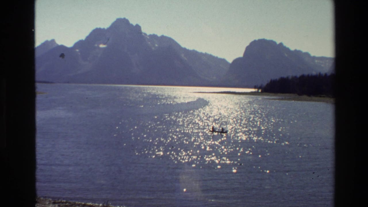 1980 Grand Teton home-movie view of a bright lake with a small canoe, dark mountain peaks, shoreline trees, and visible 8mm frame borders.