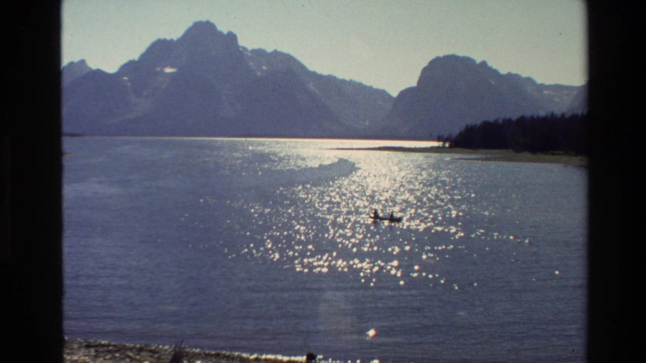 Wide lake view with canoe on reflective water, mountain peaks, and dark shoreline trees.