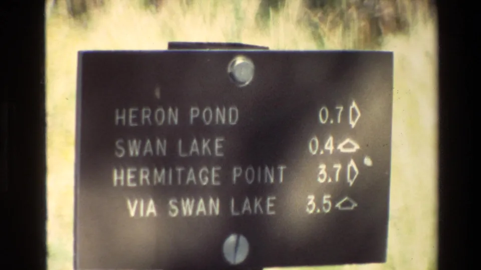 Close 1980 Grand Teton home-movie view of a black directional sign naming Heron Pond, Swan Lake, and Hermitage Point with mile distances.