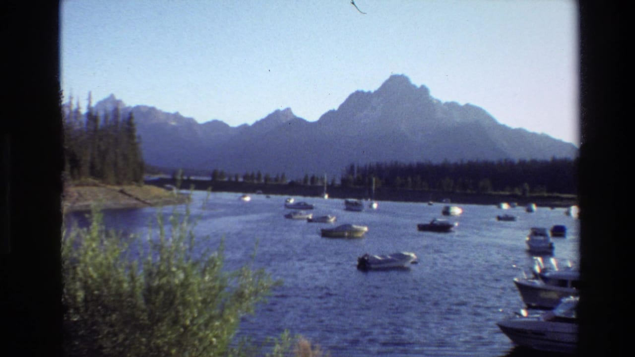 Boats spread across a lake with a wooded shoreline and high mountains behind them.