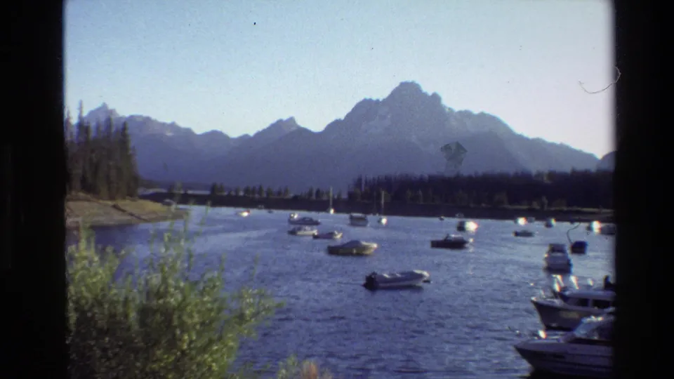 Vintage 8mm Grand Teton lake view with boats on the water, shrubs in the foreground, and mountain silhouettes beyond the shoreline.
