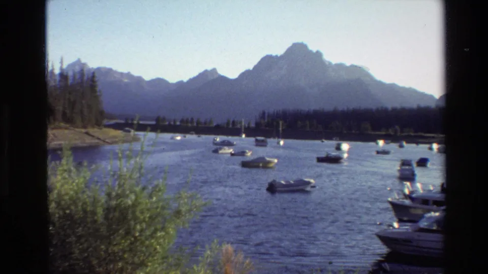 1980 Grand Teton home-movie frame showing anchored boats on blue water, a forested shore, and mountain peaks in the background.