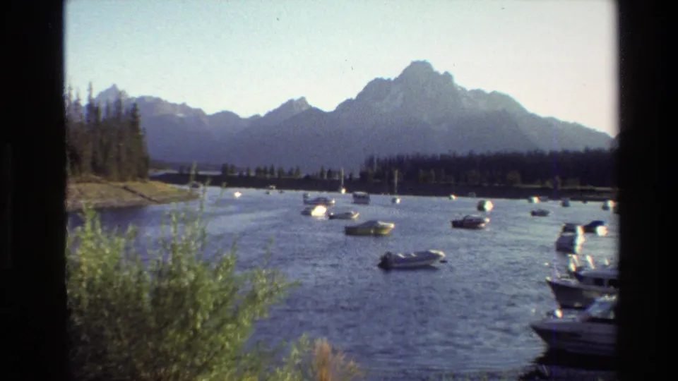 1980 home-movie frame of a Wyoming mountain lake, with boats on the water, shrubs in front, and the Grand Teton range in back.