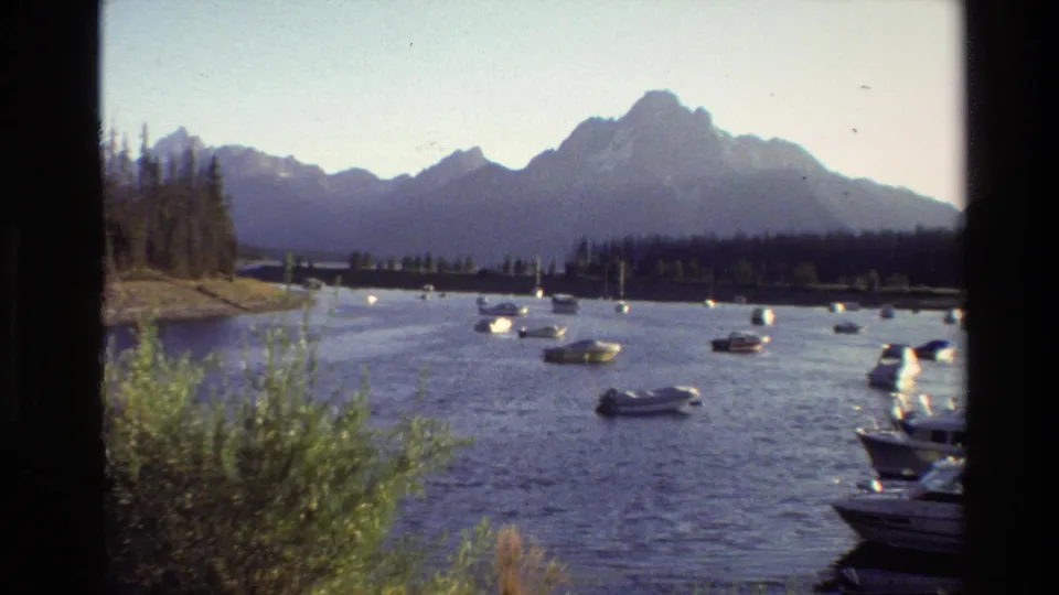 Grand Teton area lake footage from 1980, showing scattered boats, a wooded shore, foreground greenery, and distant mountain silhouettes.