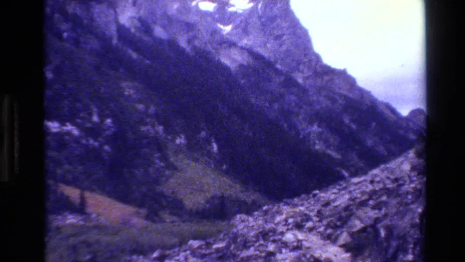 1980 8mm Grand Teton view of a rugged valley beneath jagged peaks, with evergreen slopes, rocky ground, snow patches, and a purple film cast.