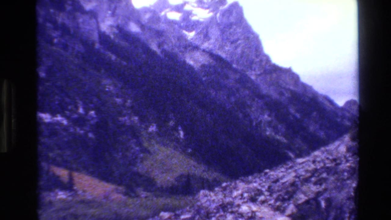 Grand Teton mountain scenery from 1980 8mm footage, showing steep forested slopes, talus, snow traces, and a bright overcast sky.