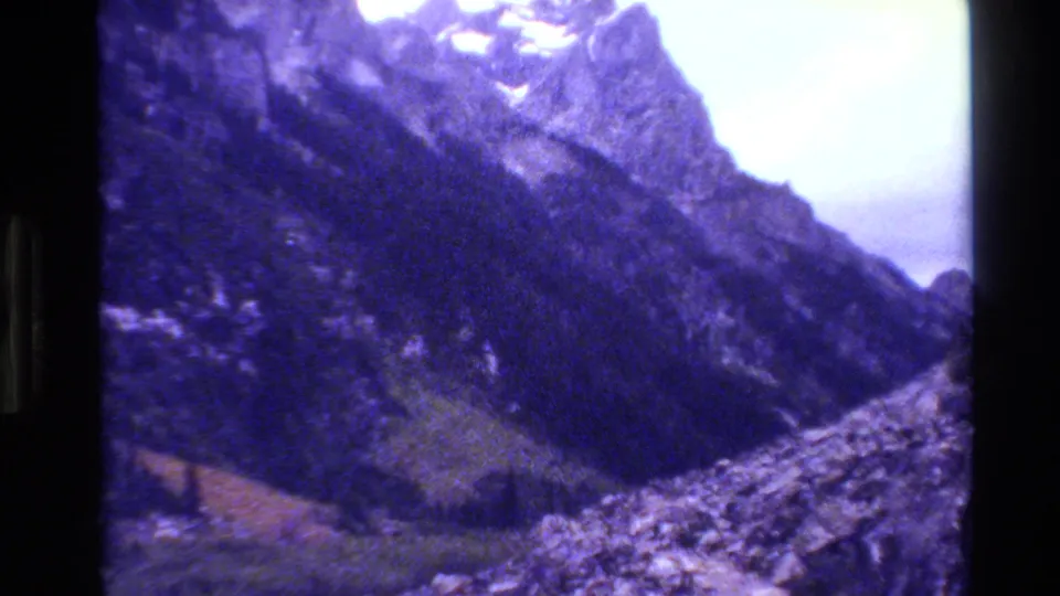 Grand Teton mountain scenery from 1980 8mm footage, showing steep forested slopes, talus, snow traces, and a bright overcast sky.