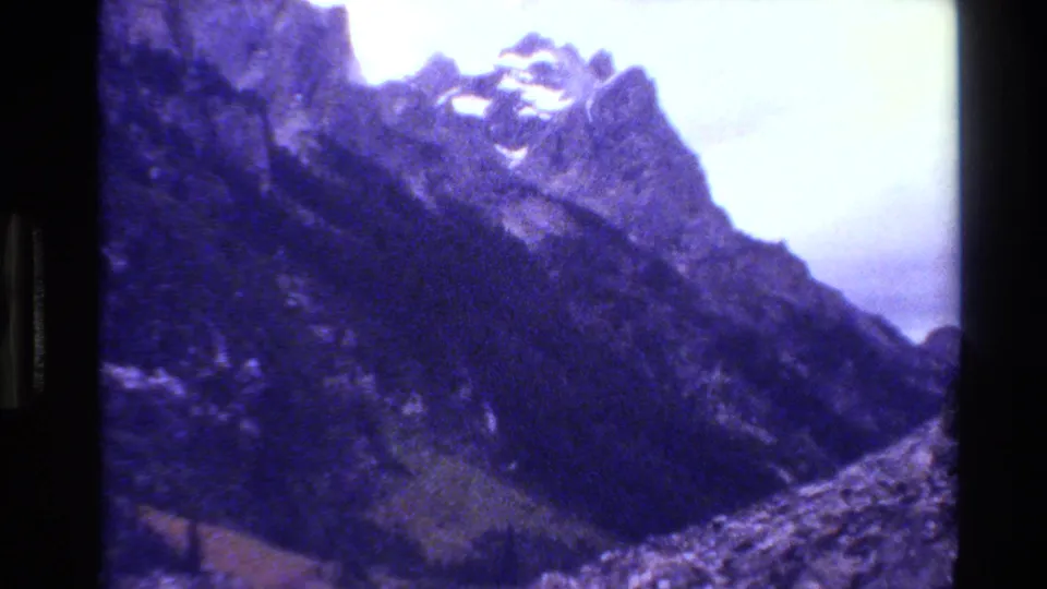 A closer 1980 Grand Teton frame shows jagged peaks, dark forested slopes, rocky valley terrain, snow remnants, and visible 8mm grain.
