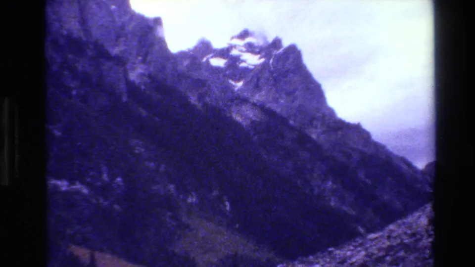 Final 1980 8mm Grand Teton still with a steep ridge, snow patches near the top, dense evergreen slope, rocky lower edge, and heavy grain.