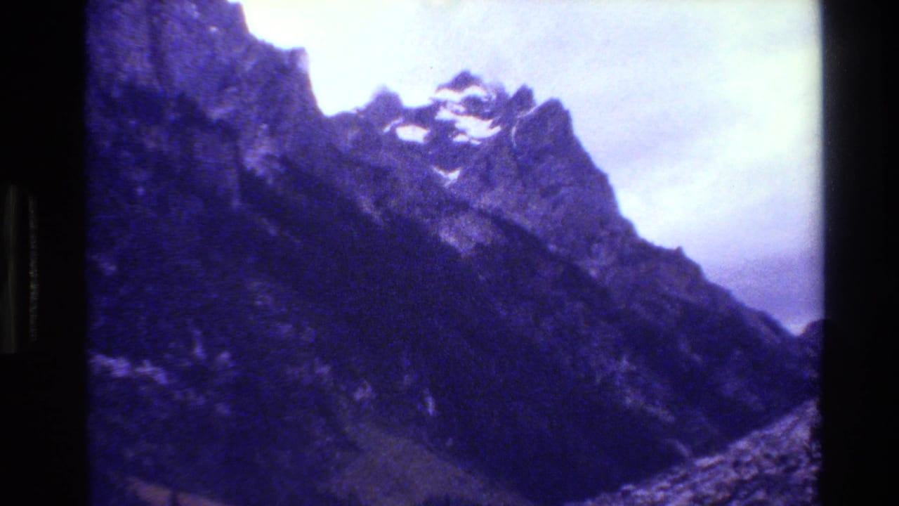 1980 8mm home-movie frame of the Grand Teton range, showing a dark rocky ridge, snow traces, and a cloudy sky with blue-purple film cast.