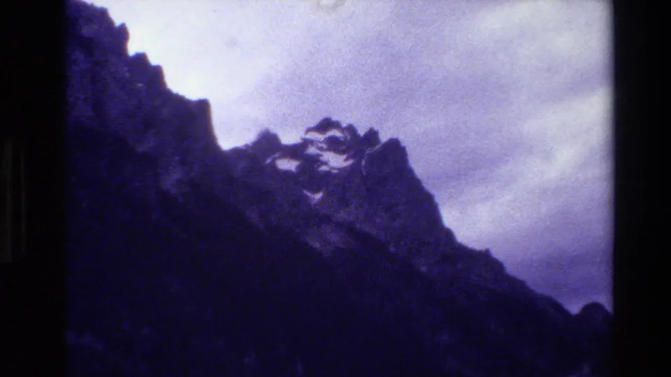 1980 Grand Teton home-movie still with rocky peaks, a dark sloping foreground, faint snow, and a washed cloudy sky.