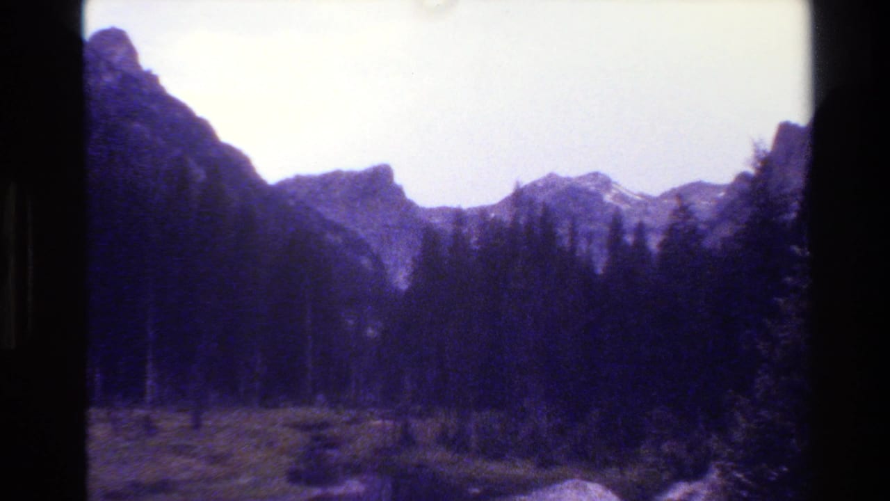 Jagged mountain skyline, evergreen trees, and meadow framed by dark film edges.