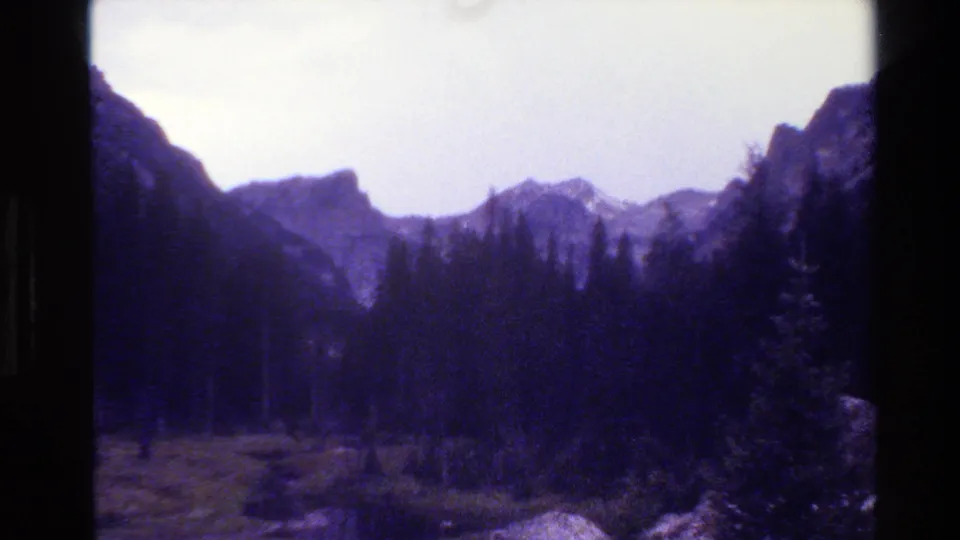 1980 Grand Teton, Wyoming frame with a forested valley, distant rocky peaks, meadow grass, and aged 8mm film texture.