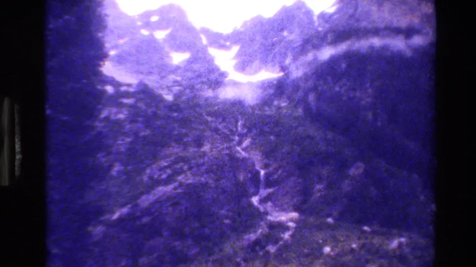 Grand Teton home-movie frame showing snow-capped peaks, rocky slopes, and a winding stream or snowmelt trace in the valley.