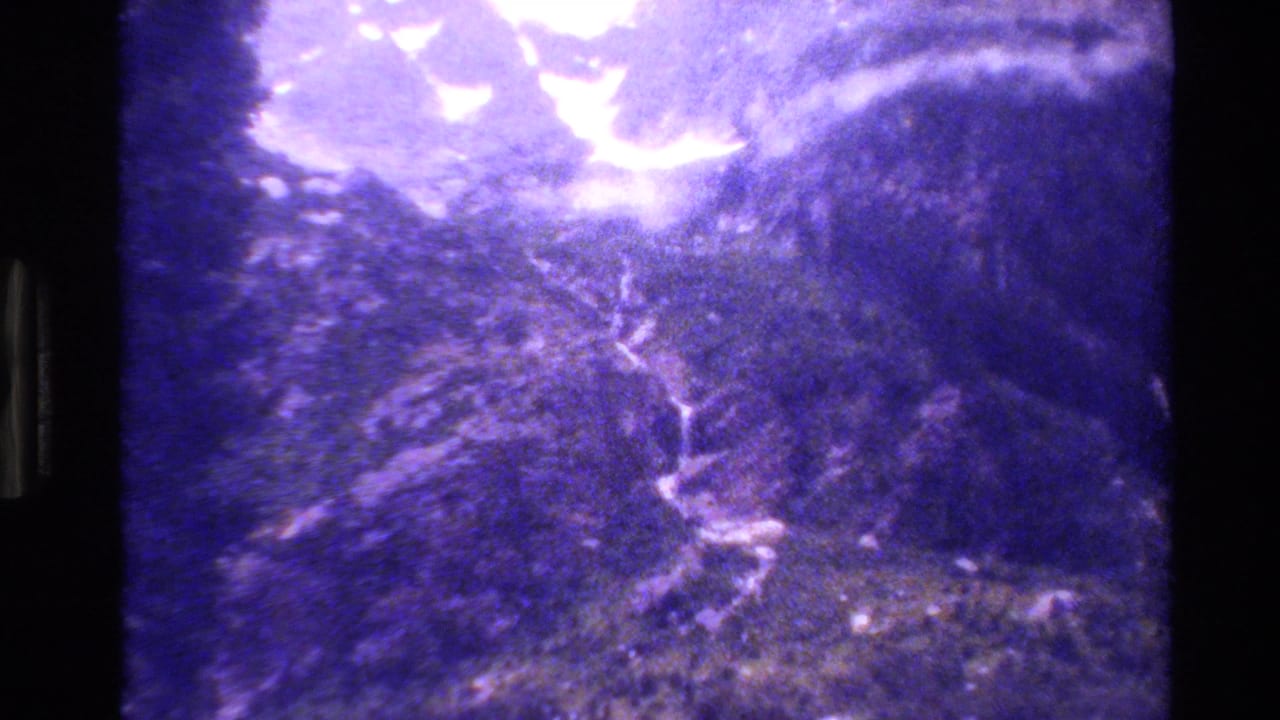 1980 Grand Teton 8mm frame showing rugged forested mountain terrain, bright overexposed sky, and a pale winding waterfall or path through the slope.